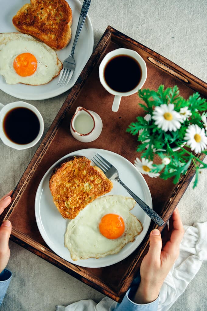 Woman holding a wooden tray with a French toast and a fried egg on a white plate and a cup of coffee, milk and flowers on the side. Another plate with french toast and an egg and another cup of coffee accompany out of the tray. 这个简单的法式吐司食谱加上辣味,让你的早晨值得醒来。它外脆里软。搭配咸的和甜的配料都很完美,但我喜欢它本来的样子。