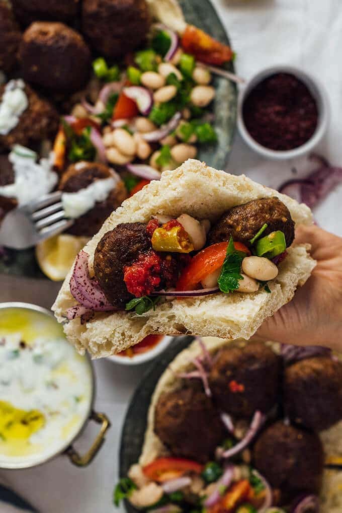 Homemade Turkish Meatballs are spicy, tender and perfectly juicy. It’s not a secret any more to make meatballs at home. #turkishfood #meatballs #meatballsrecipe #middleeasternfood #kofta #kofte 一只手拿着一个大饼三明治,里面有自制的肉丸、豆沙拉、漆树洋葱和泡菜。
