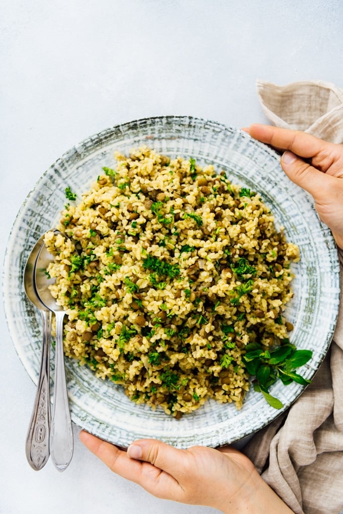 Bulgur wheat pilaf with lentils and herbs make a super healthy and tasty side dish. #pilaf #bulgur #bulgurwheat #turkishfood #bulgurrecipe 一个女人用一个大陶瓷碗盛着小麦抓饭和绿色扁豆和香草。