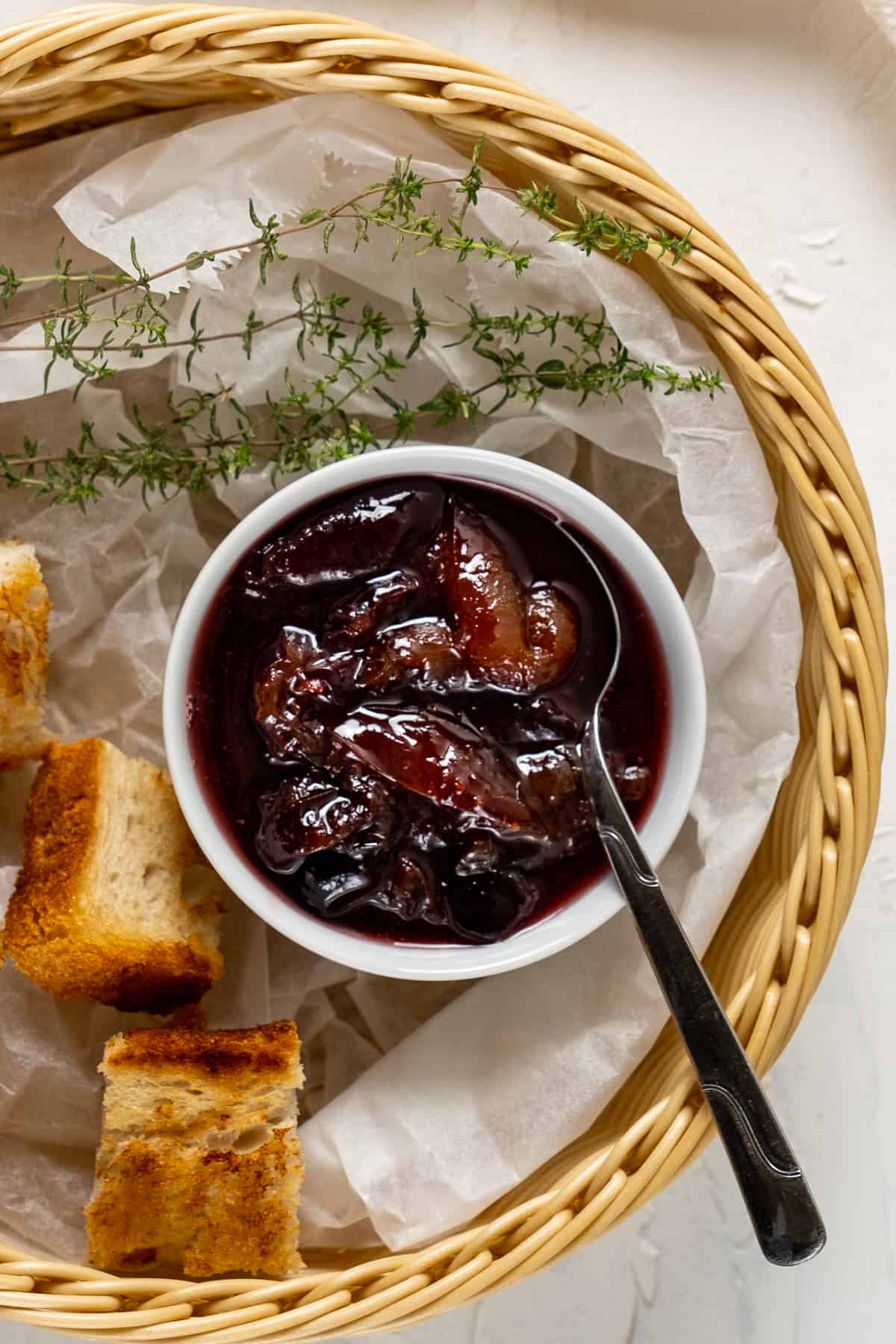 梅子酱装在一个小白碗里，里面有一把勺子，烤过的迷你面包片和新鲜的百里香都装在一个篮子里。