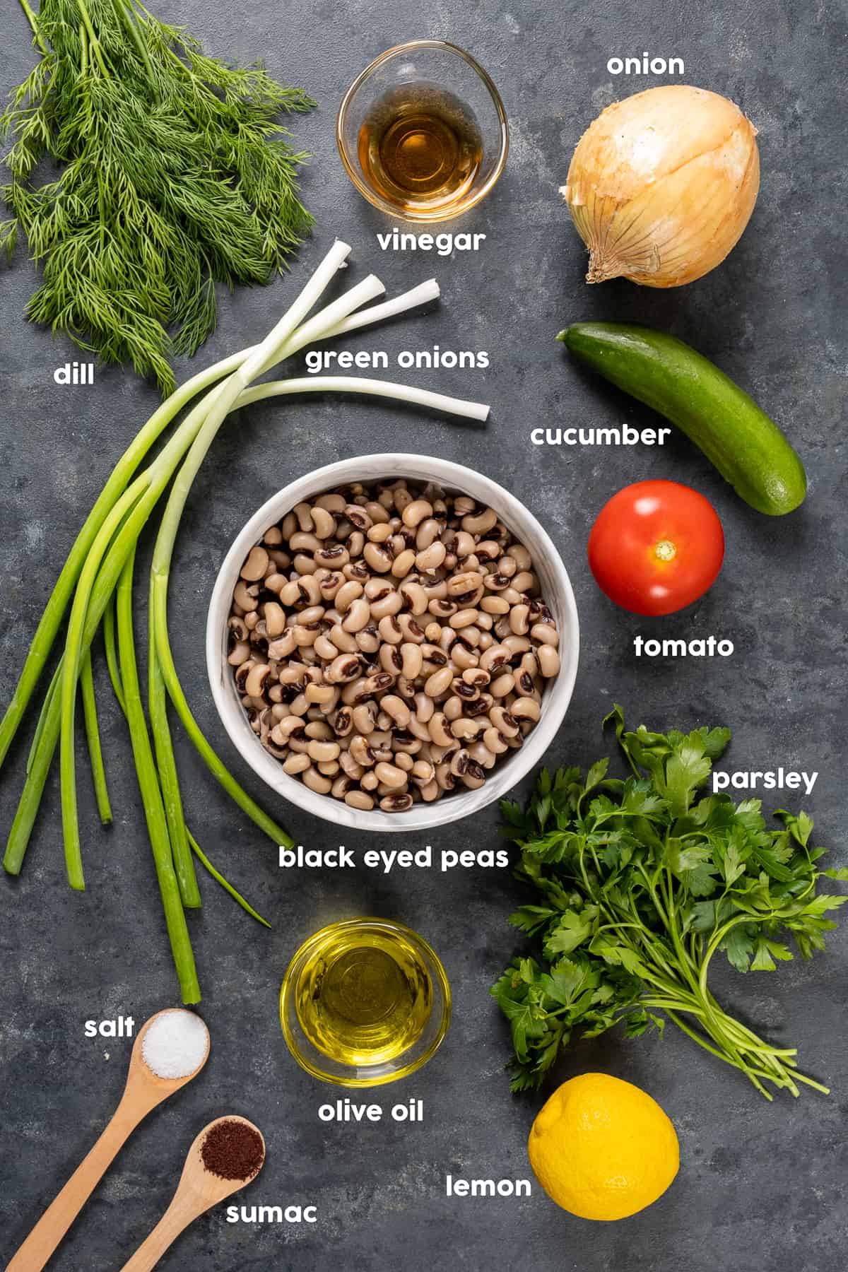 白色的碗里放着黑眼豆豆，中间是绿色的洋葱、新鲜的莳萝和欧芹、柠檬、洋葱、黄瓜、番茄、醋、盐和漆树，背景是黑色的。