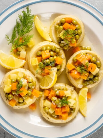 洋蓟底部塞满了土豆、豌豆和胡萝卜等蔬菜，配以新鲜的莳萝和柠檬角，放在有蓝色条纹的白色盘子里。