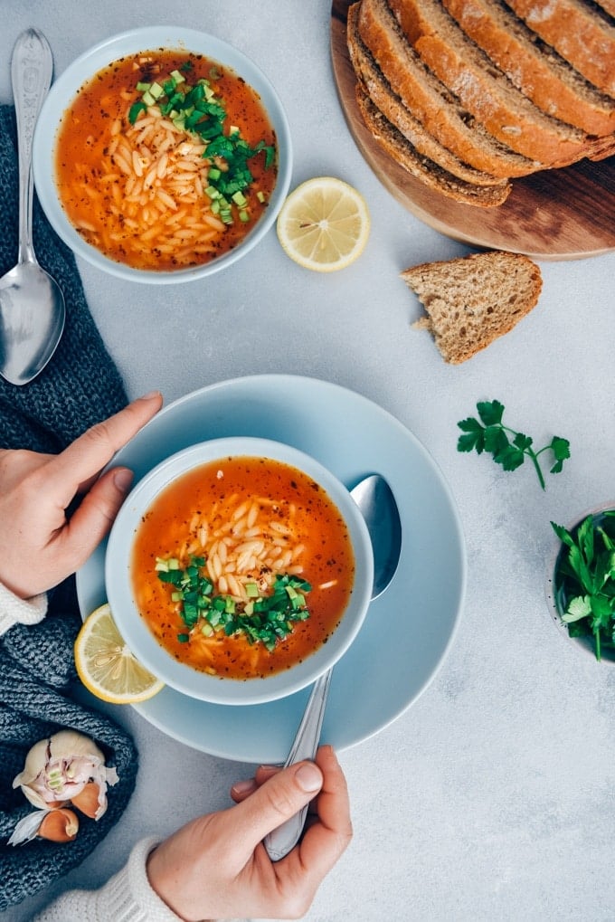 Turkish Tomato Orzo Soup Recipe packs a ton of flavor with fewer than 10 ingredients in just 20 minutes. The combination of tomatoes, lemon, parsley and orzo makes the easiest pasta soup ever. #orzosoup #wintersoup #souprecipe #turkishsoup 一名女子正在吃土耳其番茄汤,配以欧芹,盛在浅蓝色的碗里,背景是灰色的。