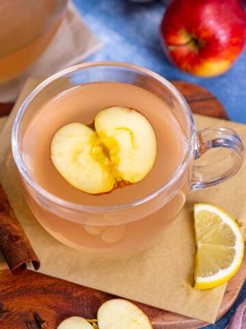 用玻璃马克杯装苹果茶，里面放一片苹果，旁边放一片柠檬和肉桂棒。