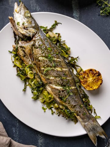 整个海鲈鱼在烤箱里烤，配上烤好的绿色蔬菜，放在白色盘子里。