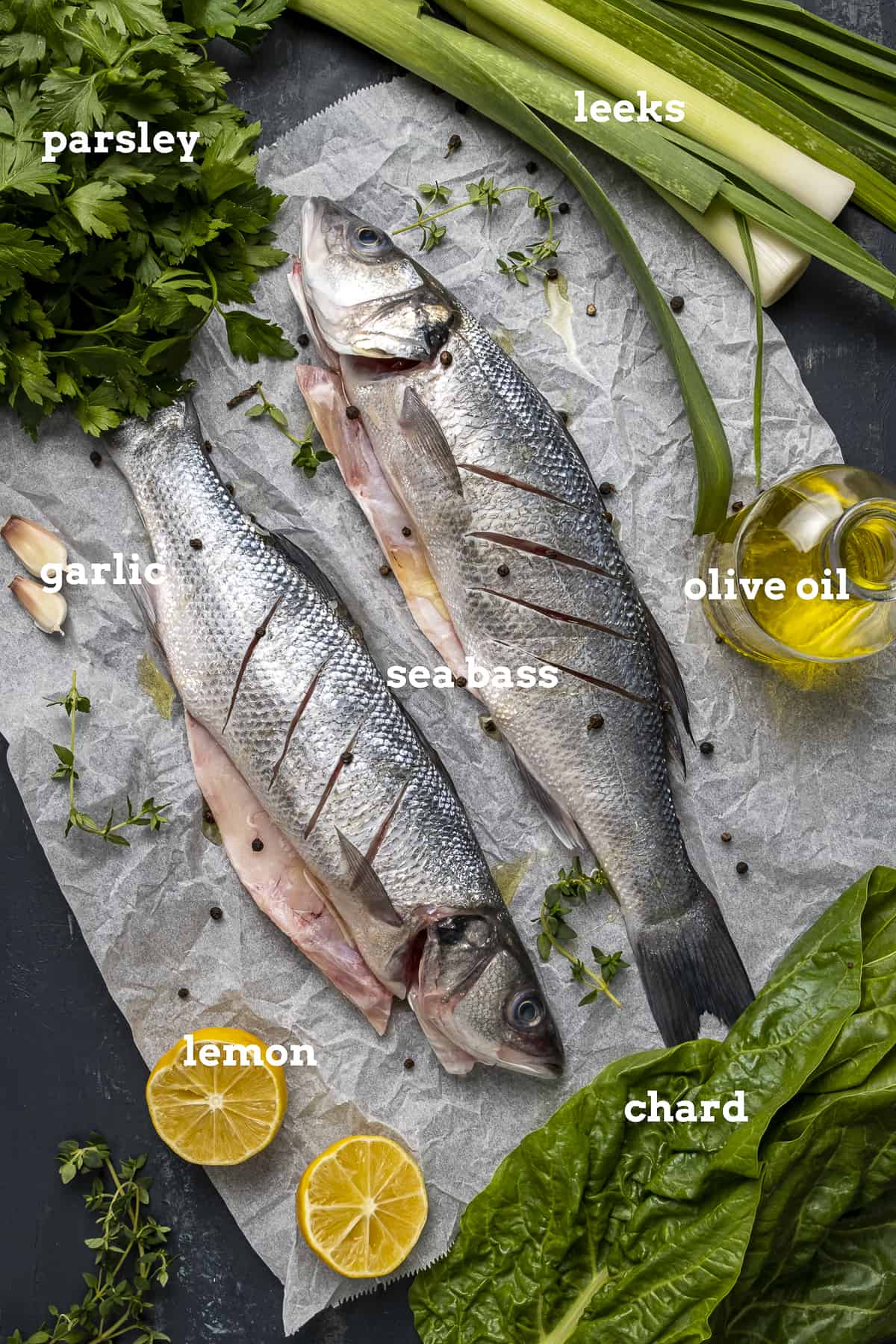 两只新鲜的海鲈鱼，蒜瓣，橄榄油，韭菜，甜菜叶，半柠檬和欧芹放在烤纸上。
