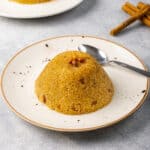 Semolina halva topped with toasted pine nuts served on a white plate with a spoon on the side.