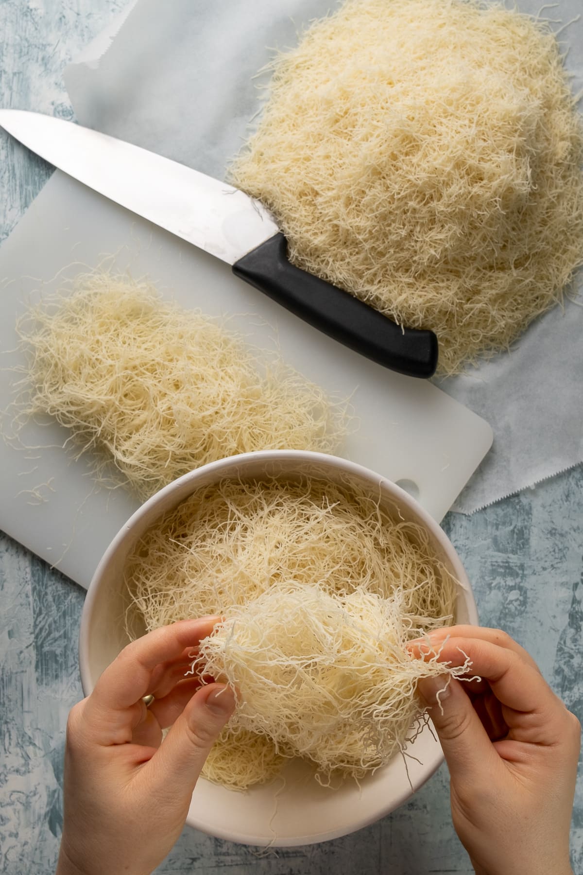 手切碎kadaif面条在碗里，更多的面条在切板和在一块烘烤纸和刀子在地面上。