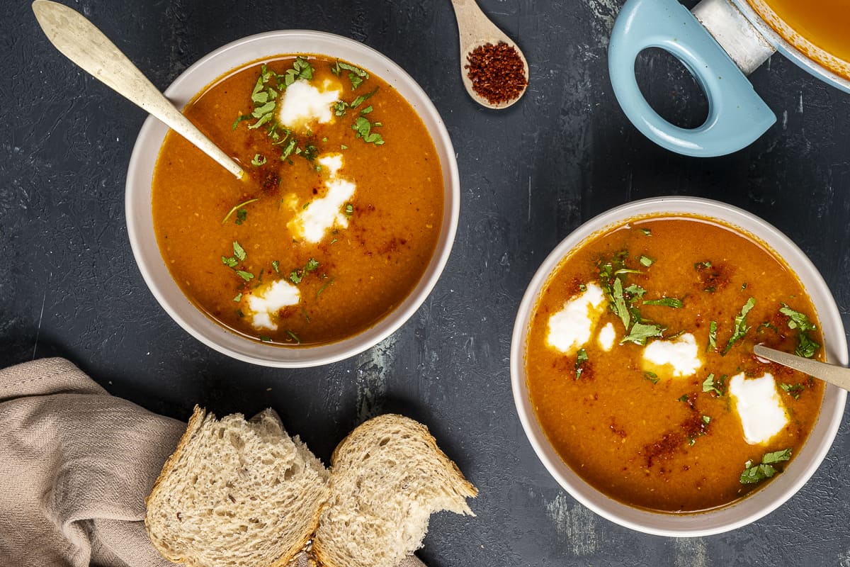 西红柿汤装在两个碗里，用酸奶和欧芹装饰，每个碗里有一个勺子。面包片和红辣椒片放在旁边。