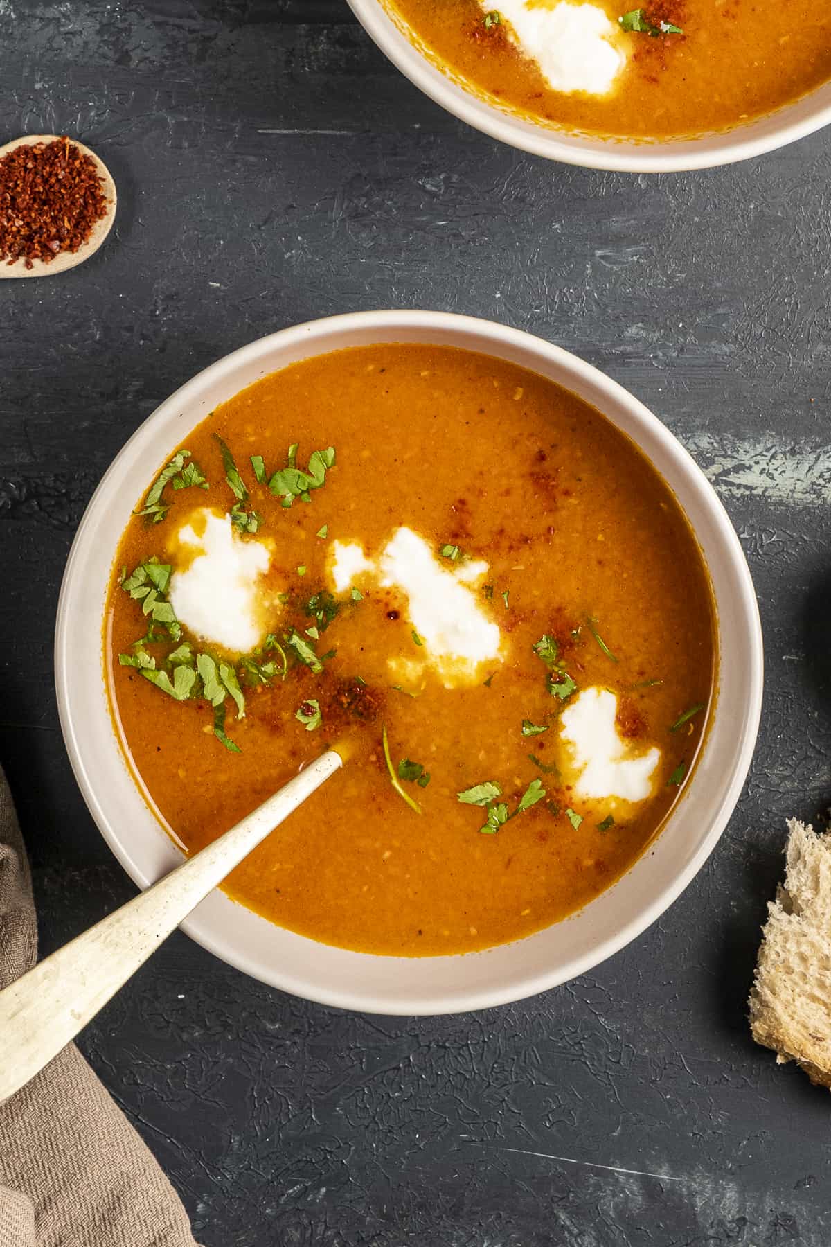 西红柿胡萝卜汤，配上酸奶、欧芹和红辣椒片，放在一个白色的碗里，里面有一个勺子。