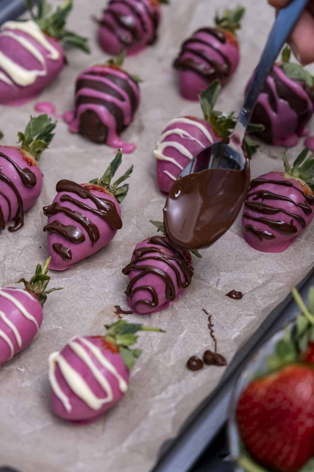 A spoon decorating pink strawberries with dark and white chocolate drizzles.