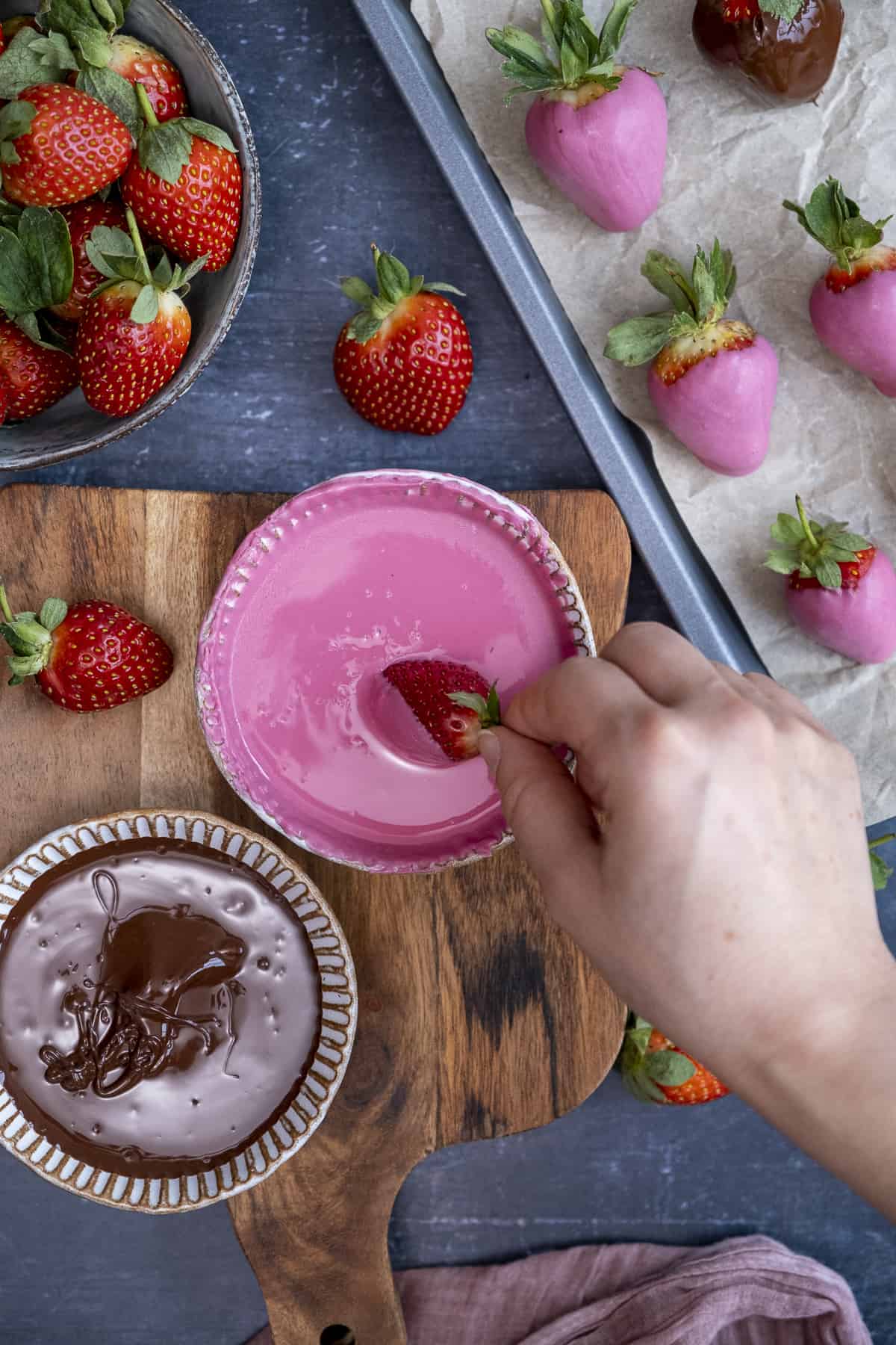 A hand dipping a strawberry into melted pink chocolate in a small white bowl, melted dark chocolate and strawberries on the side.