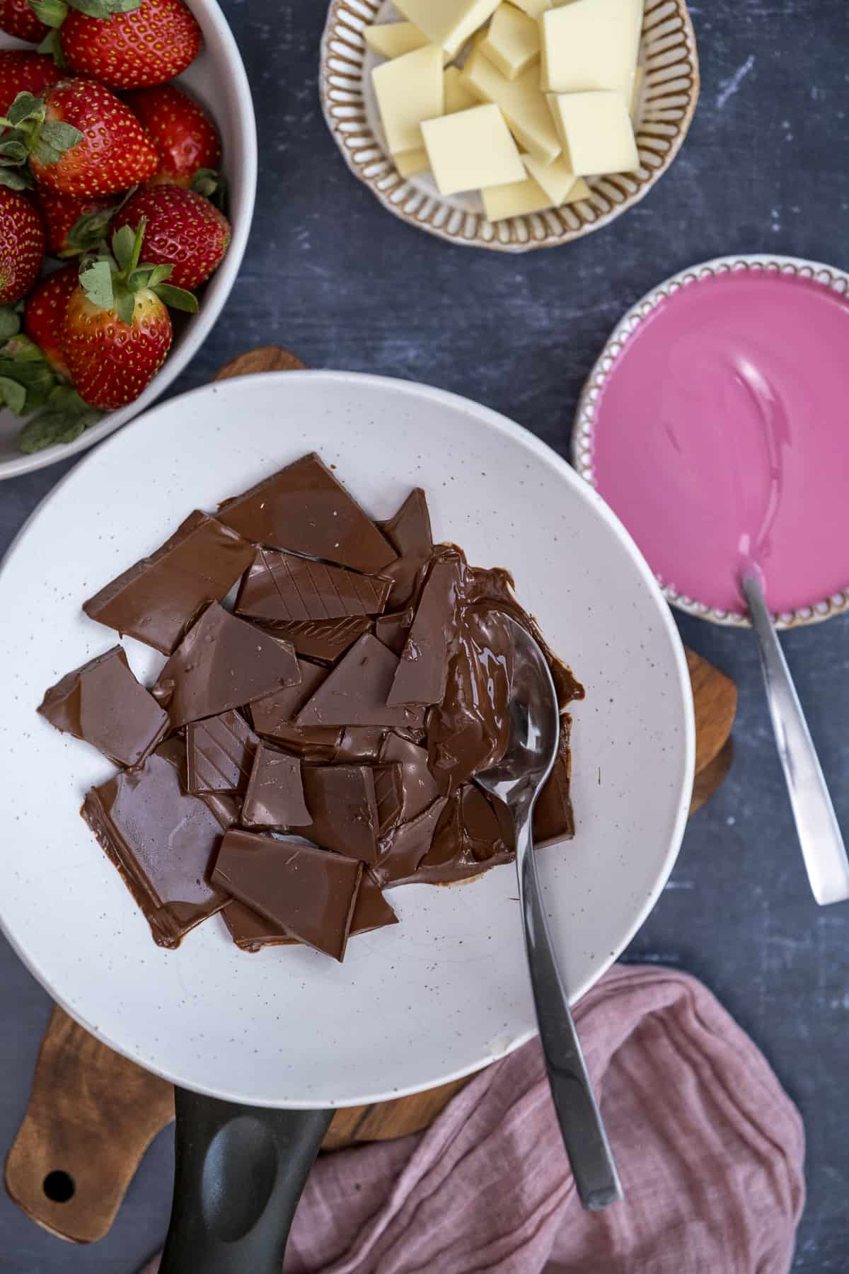 Dark chocolate melting in a white ceramic bowl over a saucepan, a spoon inside it, melted pink and white chocolate and strawberries on the side.