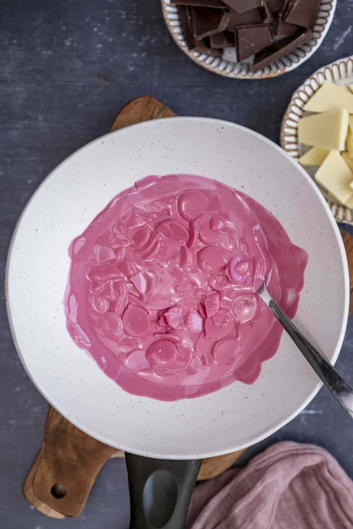 Pink candy melts melting in a white ceramic bowl over a saucepan and a spoon inside it.