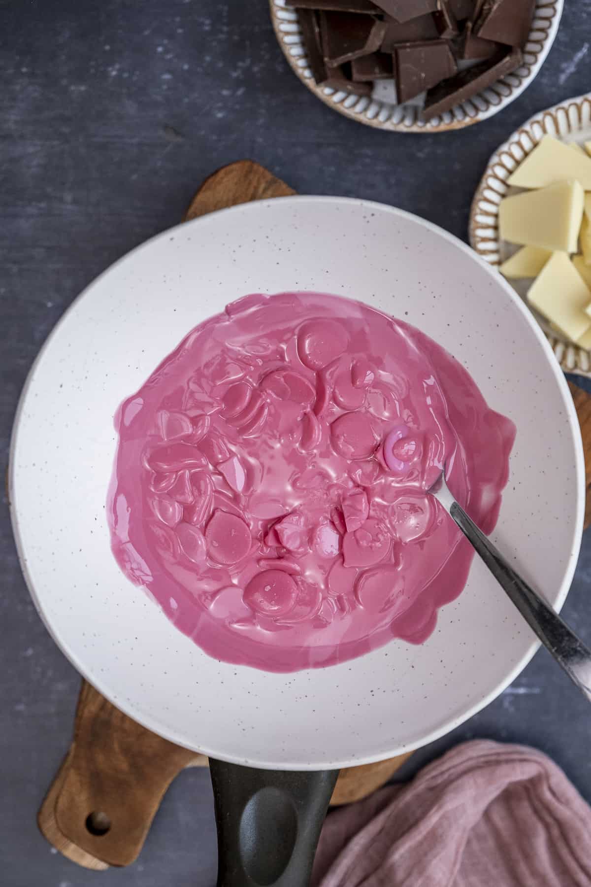 Pink candy melts melting in a white ceramic bowl over a saucepan and a spoon inside it.