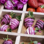 Pink chocolate strawberries in a wooden box with sections, pink roses and fresh strawberries behind them.