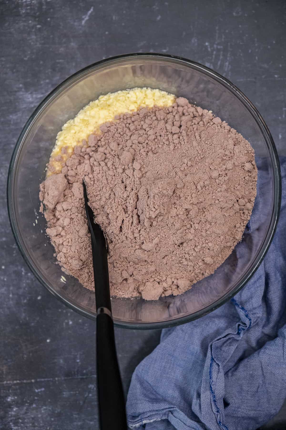红色丝绒蛋糕粉和鸡蛋和黄油的混合物在一个玻璃搅拌碗里,里面有一个黑色的锅铲。