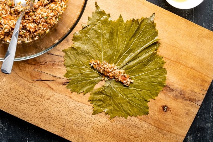 Some rice filling mixture is put on a vine leaf on a wooden board, filling bowl on the side.