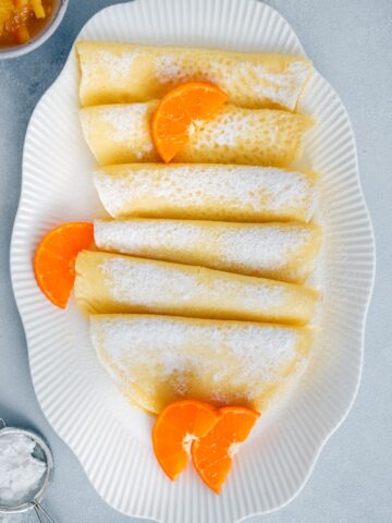 无麸质可丽饼装在白色的椭圆形盘子里，用橘子片和糖粉装饰。