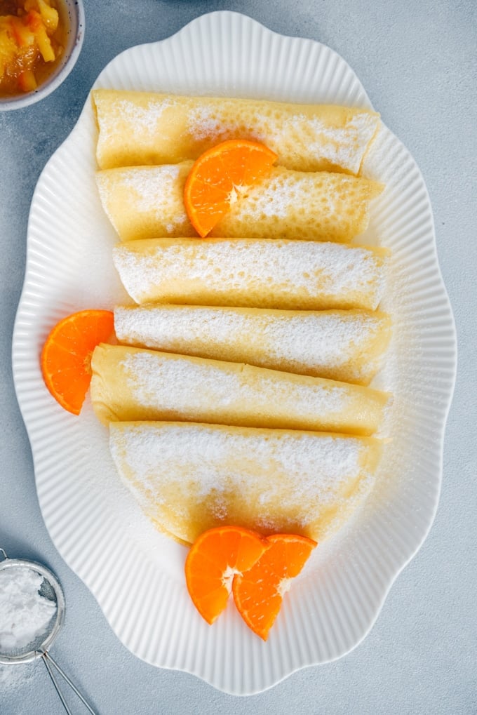 Wonderfully thin gluten free crepes made with rice flour are no different from regular crepes! So easy to make with only 4 simple ingredients. Perfect for several filling options! #glutenfreebreakfast #glutenfreecrepes #riceflourrecipes 无谷蛋白法式薄饼在白色椭圆盘,点缀以橙片和糖粉。