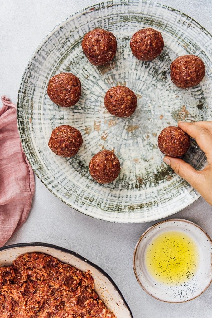 Homemade Turkish Meatballs are spicy, tender and perfectly juicy. It’s not a secret any more to make meatballs at home. #turkishfood #meatballs #meatballsrecipe #middleeasternfood #kofta #kofte 一只手把生的自制鱼丸放在陶瓷盘子上,旁边的小碗里放着橄榄油。