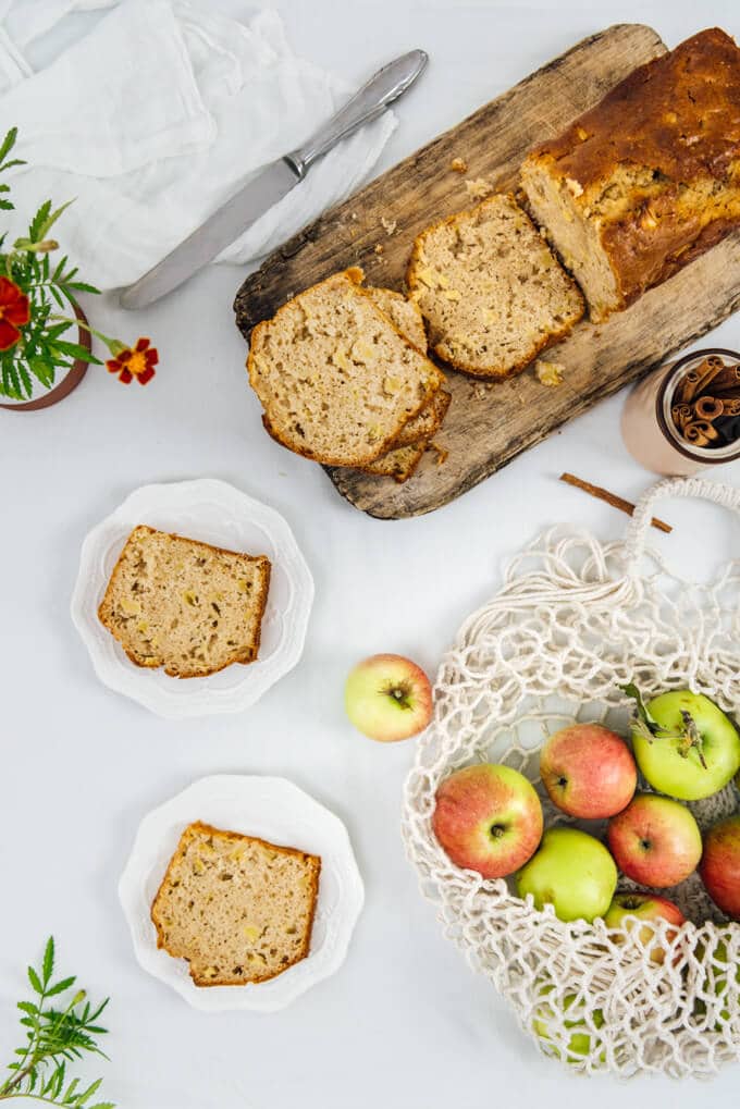 厚实的苹果肉桂面包切在木板上，装在两个白色盘子里，苹果装在一个细绳袋里。