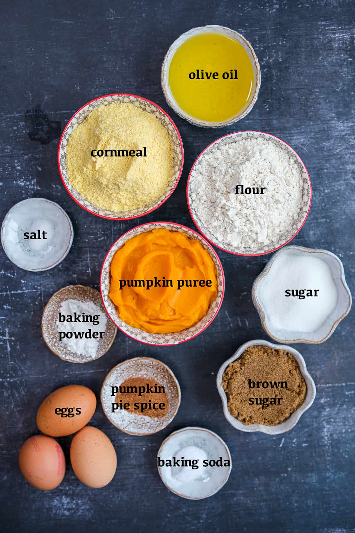 南瓜泥、玉米粉、面粉、橄榄油、红糖、砂糖、鸡蛋、南瓜香料、盐、小苏打和泡打粉都放在不同的碗里，背景是深色的。
