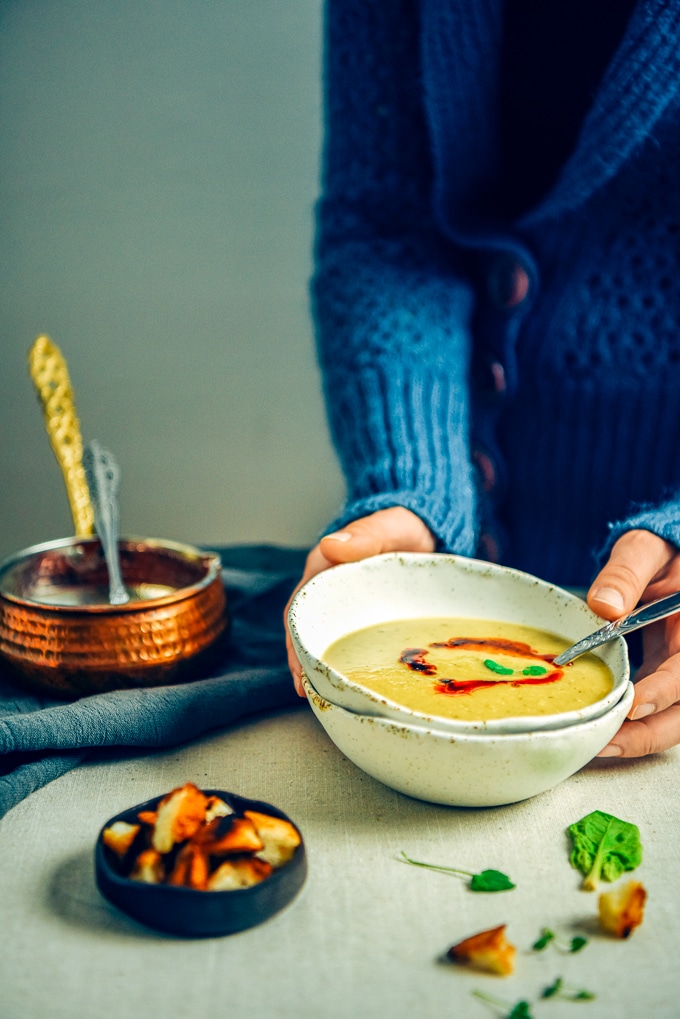 一个穿着蓝色开襟羊毛衫的女人端着一碗土耳其扁豆汤。铜制调味锅，配香草和油炸面包丁。