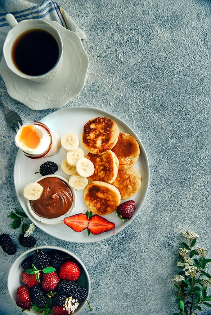 早餐，一杯茶配酸奶煎饼、浆果、香蕉片、巧克力和一个白盘子煮熟的鸡蛋