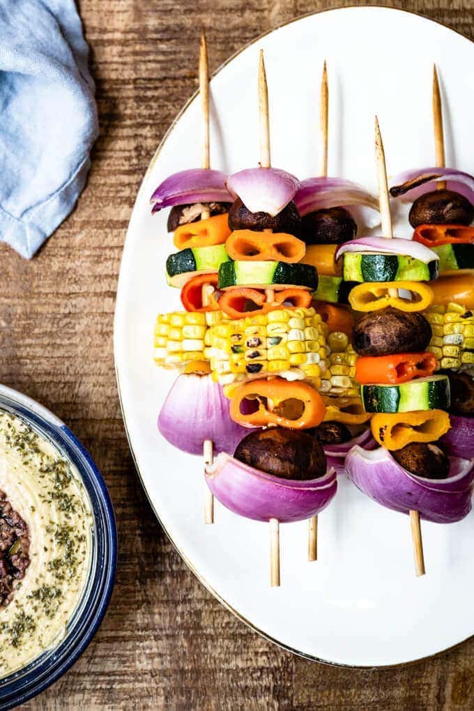 Veggie Kabobs with a simple tangy marinade are so tasty and healthy. So vibrant and packed with flavors, these kabobs make a a great side dish to any meal or a perfect summer appetizer. #vegetarian #kabobs #summerparty #partyfood #meatless #appetizer #ad @sabradips 彩色的蔬菜串放在白色的盘子里,配上蓝色的餐巾和鹰嘴豆泥。