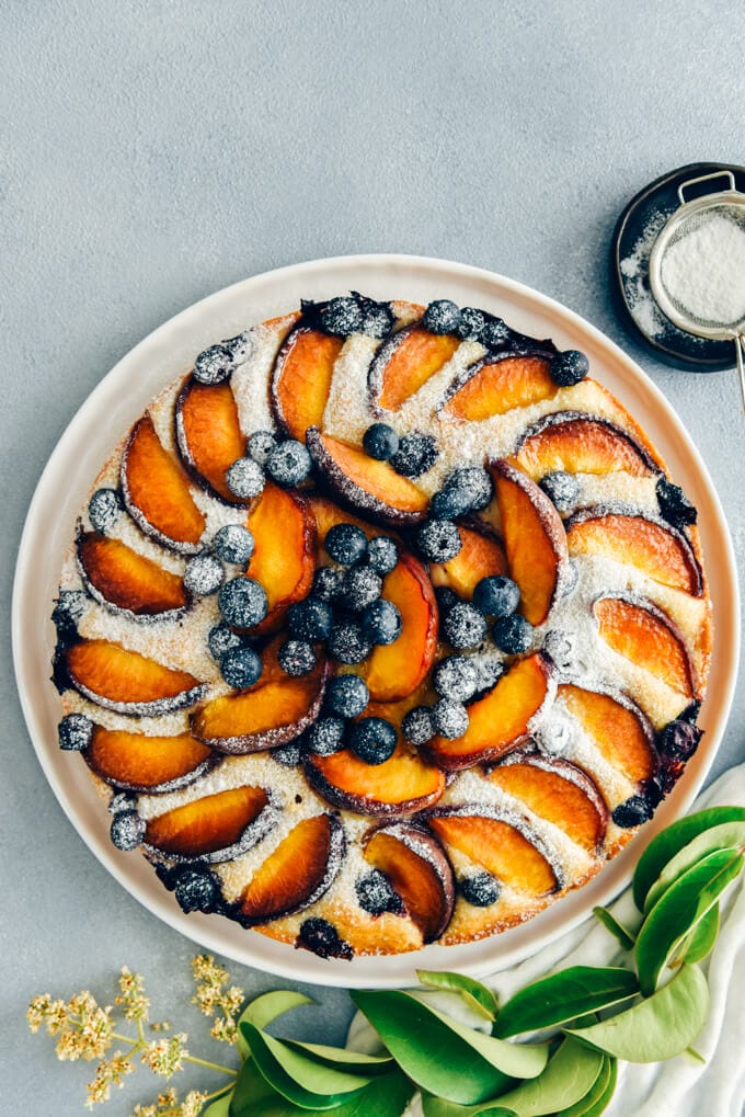 This Blueberry Peach Coffee Cake is a luscious cake that is sure to be a crowd pleaser. Made with olive oil and kefir, this simple cake is moist, tender and so light. #peachcake #peachcoffeecake #peachrecipe #coffeecake #kefircake #kefirrecipes 白盘子上撒着糖粉的蓝莓桃咖啡蛋糕