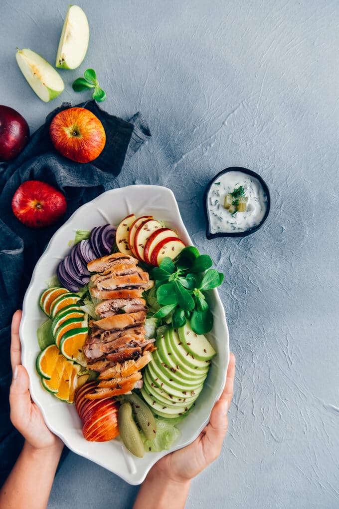 Chicken Salad with Apples is loaded with crisp apples, juicy oranges and red onions. The mouthwatering yogurt dressing with pickles makes this fall salad a big hit. #chickensalad #fallsalad #chickenapplesalad 手放在沙拉碗里,里面有鸡肉沙拉、苹果、橘子片和香草。