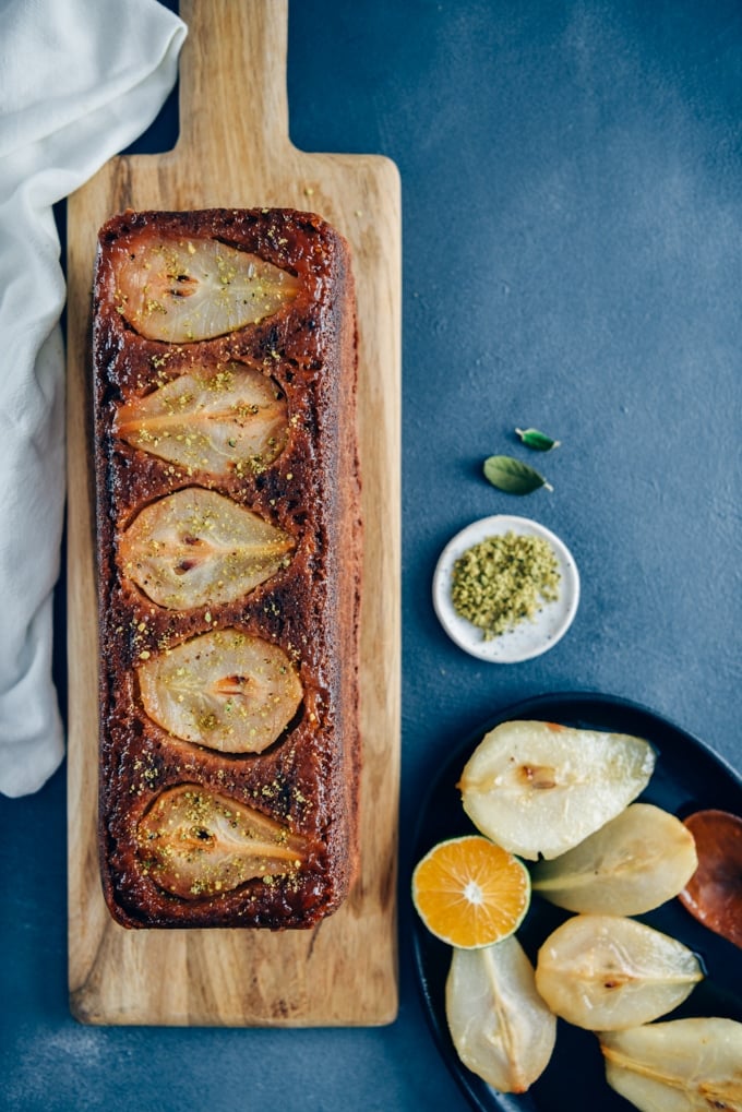 This fantastic Caramelized Pear Bread will become your ultimate fall bread recipe because it’s irresistible with the impressive pears on the top and all the flavors packed in it. #pearrecipe #pearbread #fallrecipe 木板上的梨面包,旁边的铁锅里放着焦糖梨和一块橙子。
