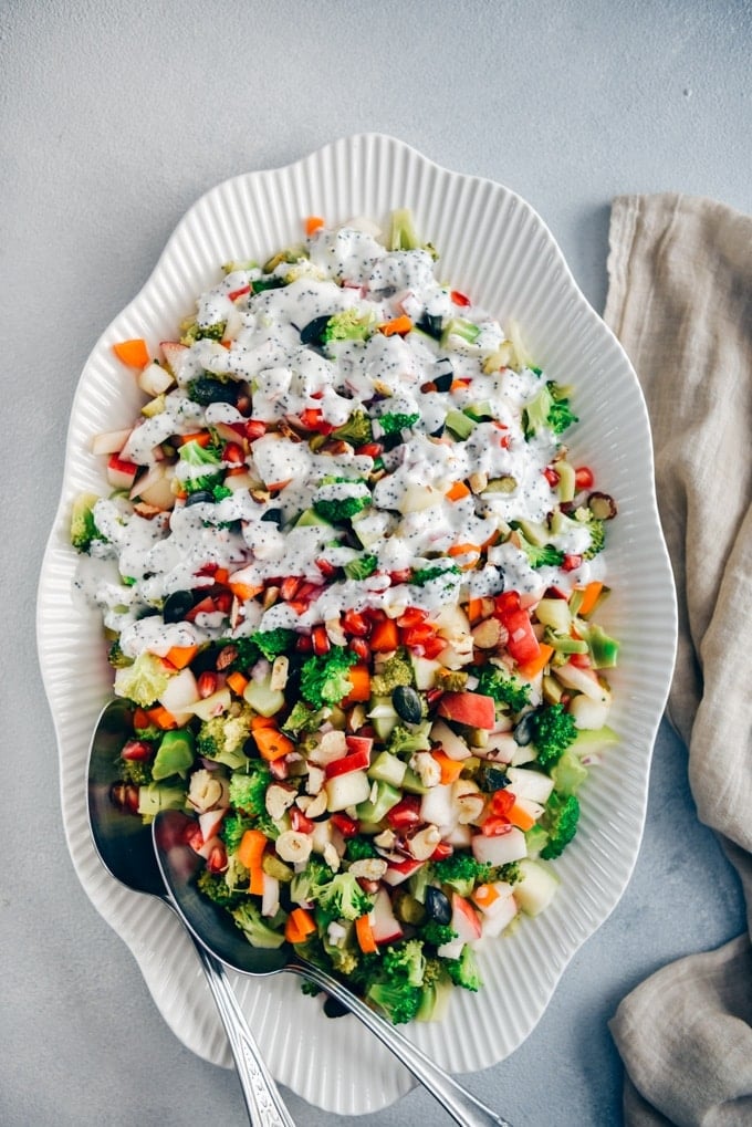 Nutrition packed colorful Broccoli Apple Salad. Ready in 10 minutes, including the creamy and oh-so-yummy yogurt dressing! #applesalad #broccoliapplesalad #holidaysidedish 西兰花苹果沙拉,胡萝卜,坚果和南瓜籽淋上罂粟籽酸奶酱,从俯视图拍摄在一个白色椭圆形碗里。