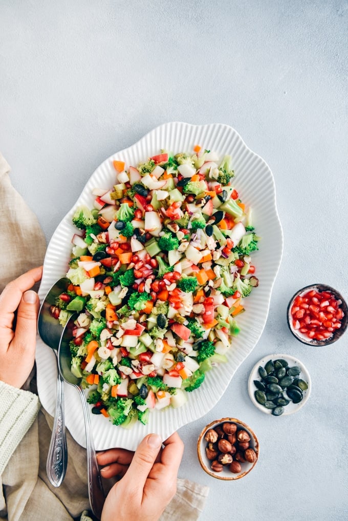 Nutrition packed colorful Broccoli Apple Salad. Ready in 10 minutes, including the creamy and oh-so-yummy yogurt dressing! #applesalad #broccoliapplesalad #holidaysidedish 手拿花椰菜,苹果沙拉,胡萝卜,坚果,南瓜子和石榴皮在一个椭圆形的碗。