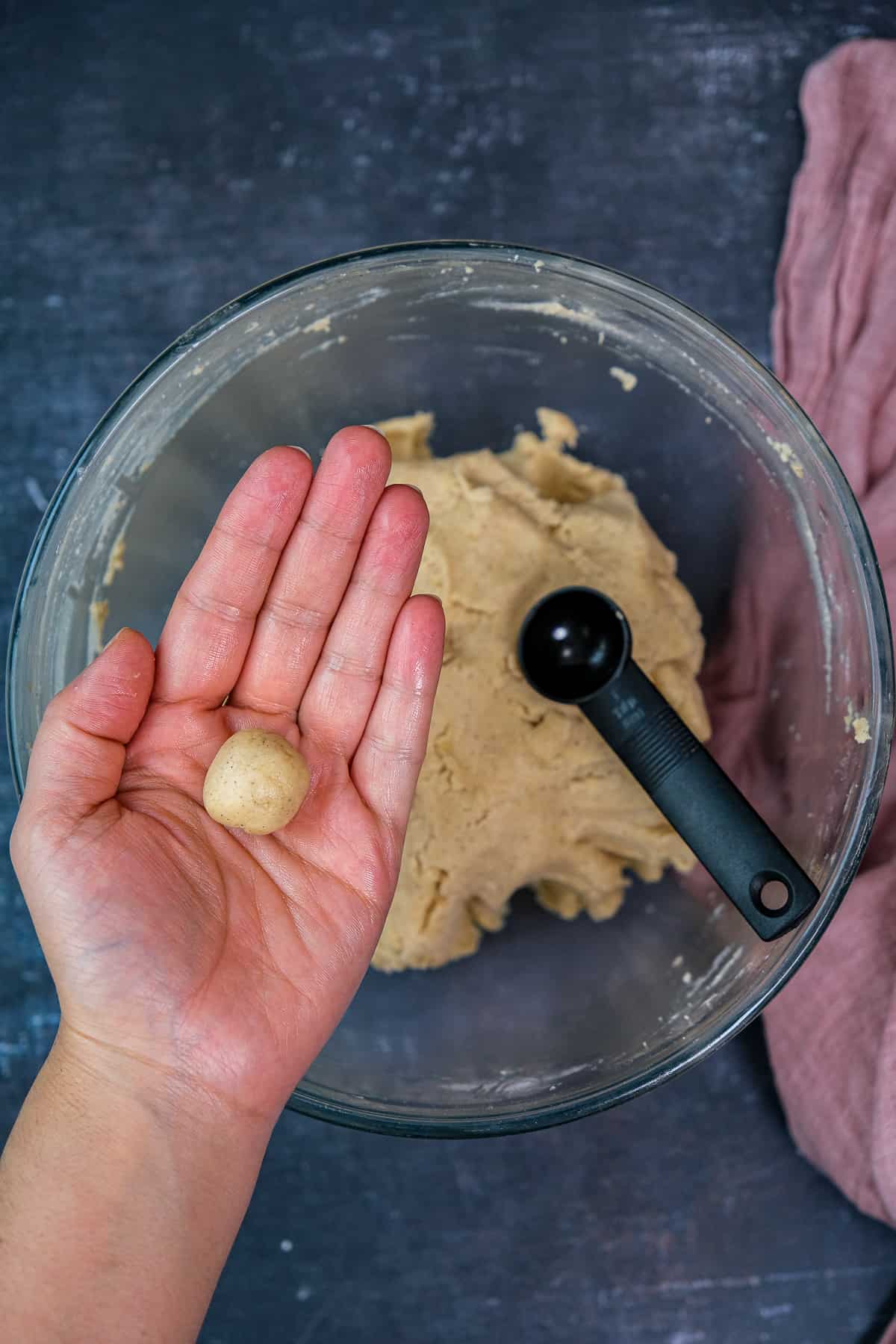 一只手拿着一个迷你曲奇面团球，剩下的面团和勺子放在下面的搅拌碗里。