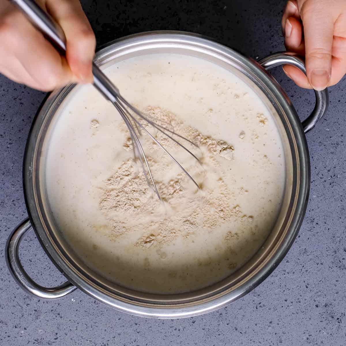 Hands whisking milk, sugar and salep powder with a hand whisk in a pan.