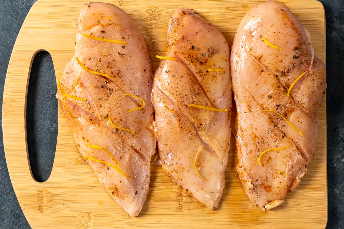 三个鸡胸肉，每个鸡胸肉上都有细缝，用香料和柠檬皮调味，放在木板上。