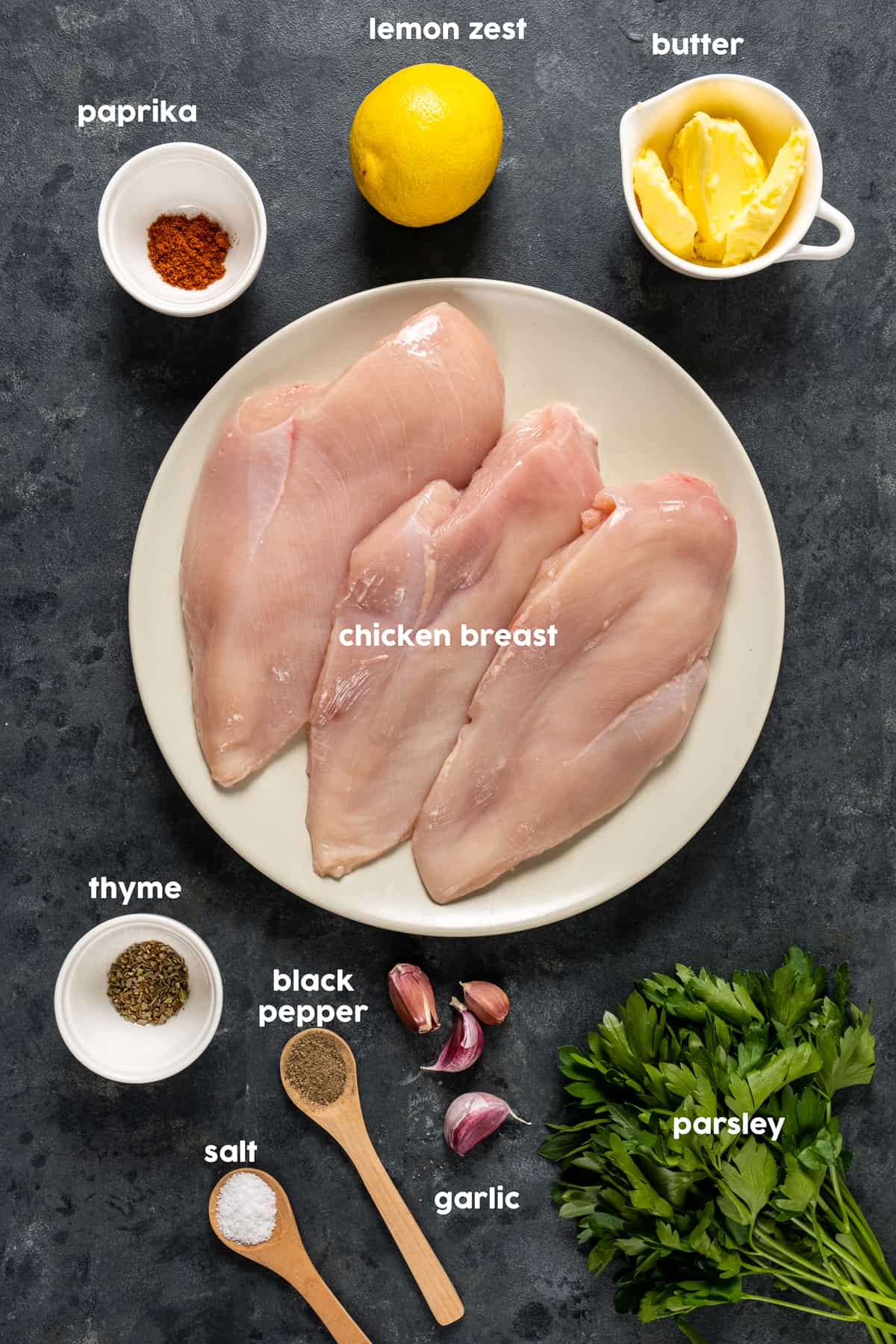 白色盘子里放着鸡胸肉,黑色背景上放着百里香、黑胡椒、蒜瓣、欧芹、黄油、柠檬和辣椒粉。
