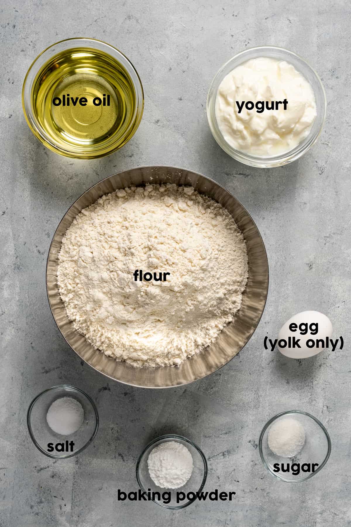 面粉、橄榄油、酸奶、盐、糖、发酵粉分别放在碗里，背景是灰色的，旁边放一个鸡蛋。