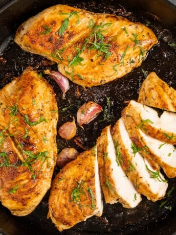 在铸铁锅里煮熟的鸡胸肉，其中一块被切成薄片，用欧芹和未剥皮的蒜瓣装饰。