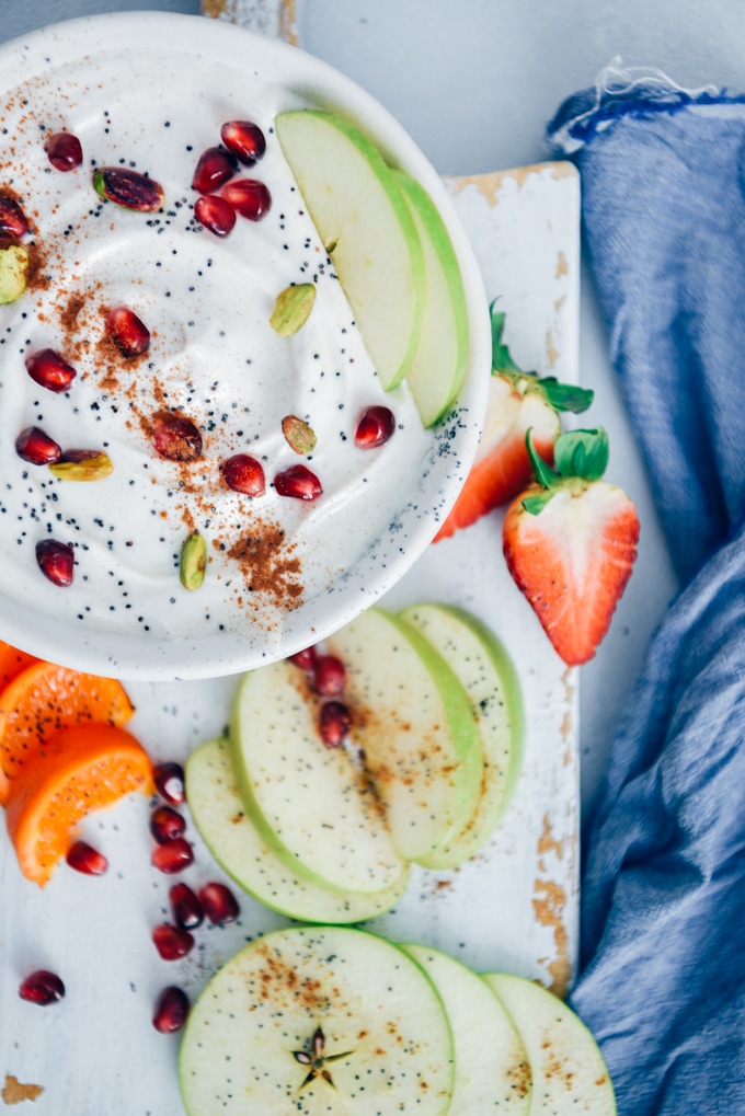 Yogurt Fruit Dip is ridiculously easy to make and always a hit. I love bringing it to parties along with a fruit platter. #snackrecipe #yogurtfruitdip #fruitdip #healthysnack 用罂粟籽、石榴籽、开心果和肉桂装饰的酸奶水果蘸酱,盛在白色的陶瓷碗里,放在白色的木板上。