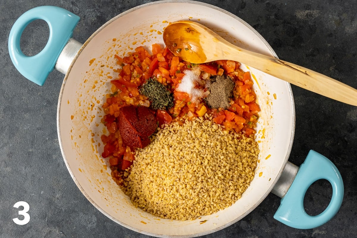 在一个白色的平底锅里放入碎gur，西红柿，番茄酱，香料，洋葱和辣椒，里面放一个木勺。