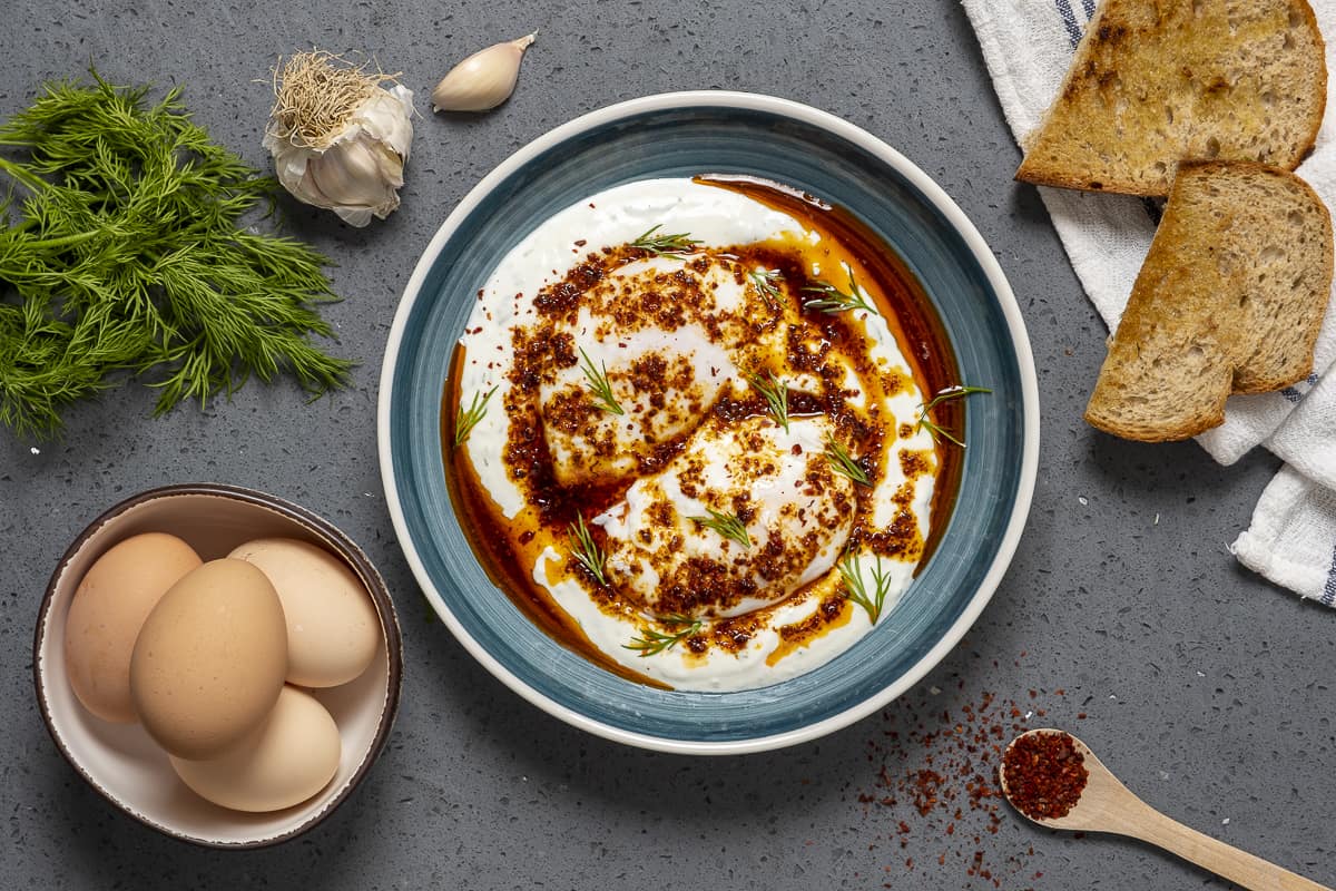 土耳其风格的荷包蛋放在蓝色的碗里，一束新鲜的莳萝，带壳的鸡蛋放在碗里，大蒜瓣，红辣椒片和烤面包在旁边。