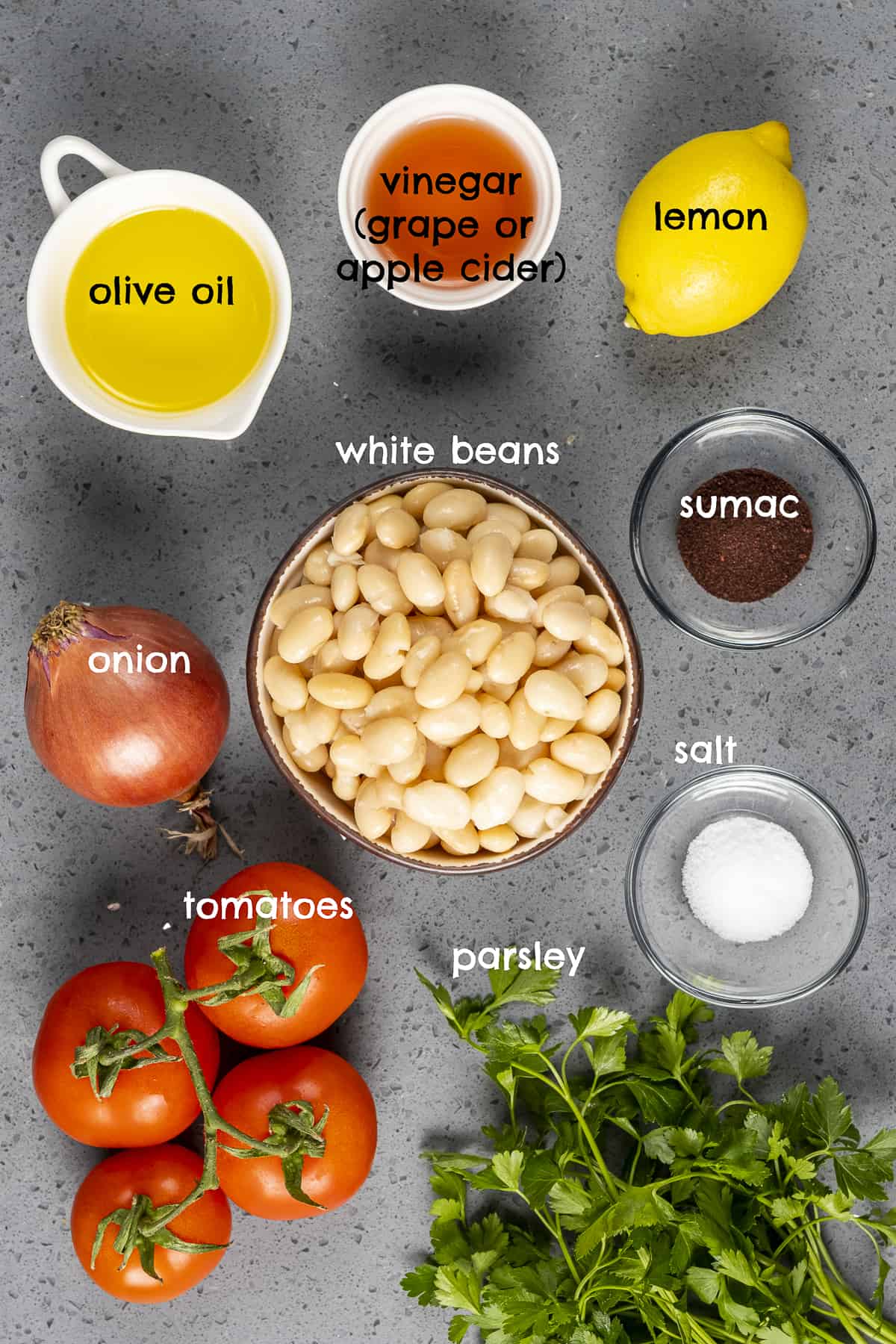 煮熟的白豆，西红柿，橄榄油，欧芹，柠檬，洋葱，漆树和盐在灰色的背景上。
