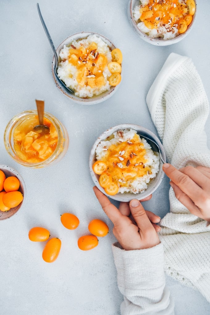 Do you have leftover rice? Turn it into a wonderful breakfast next morning by making this Sweet Rice Porridge Recipe! Don’t have cooked rice? So easy to make it from scratch too! #riceporridge #porridgerecipe 一名女子正从白色陶瓷碗里喝着盛有橘子酱、杏仁、肉桂和金桔的米粥。