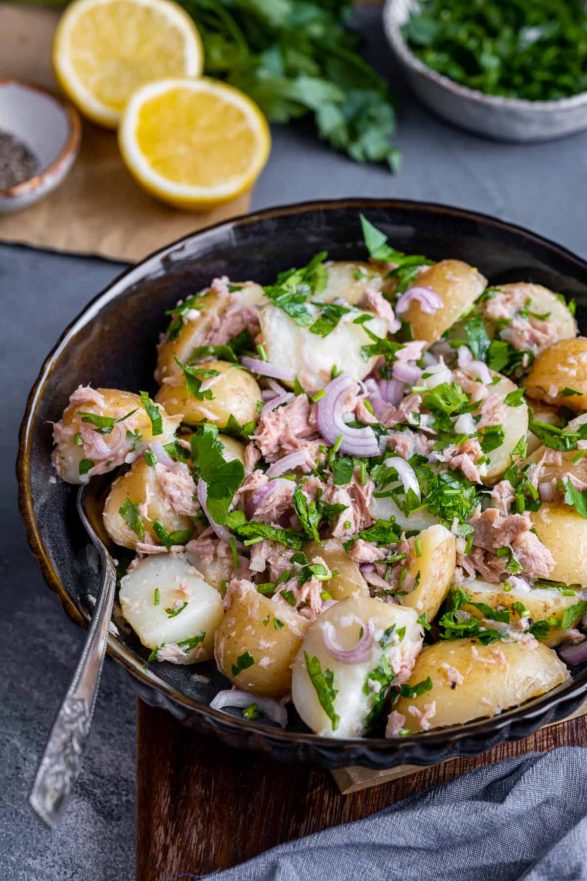土豆沙拉，金枪鱼，洋葱和欧芹放在一个黑色的碗里，里面是叉子，后面是柠檬和欧芹。