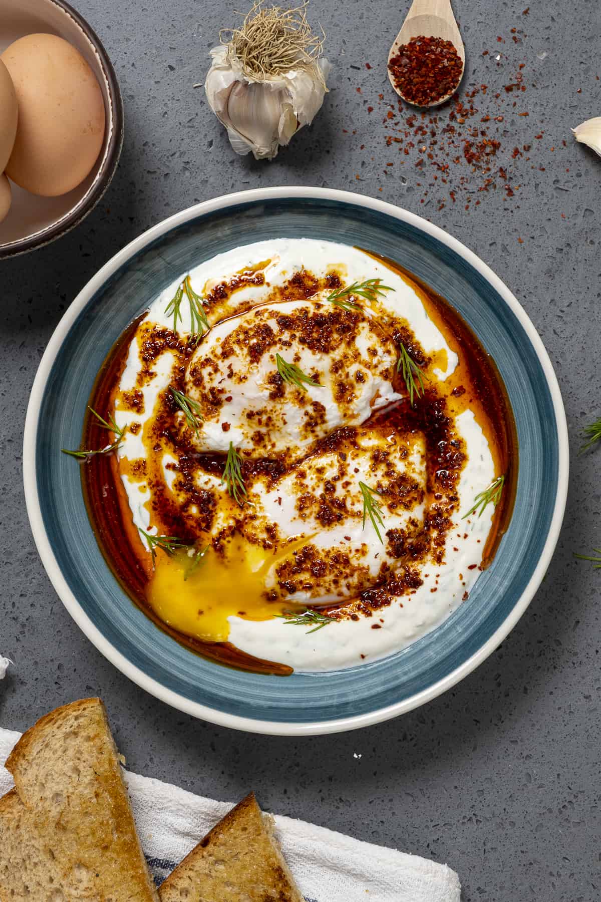 希腊酸奶上的水煮蛋，配上香料黄油酱，放在一个蓝色的碗里。蒜瓣，烤面包，胡椒片和带壳的鸡蛋放在旁边。