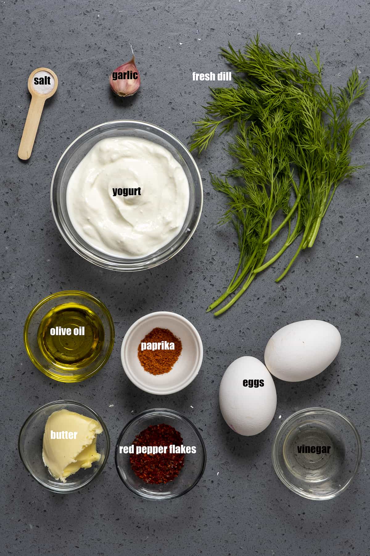 鸡蛋，希腊酸奶，新鲜的莳萝，黄油，橄榄油，红辣椒片，辣椒粉，蒜瓣，盐在一茶匙的深色背景上。