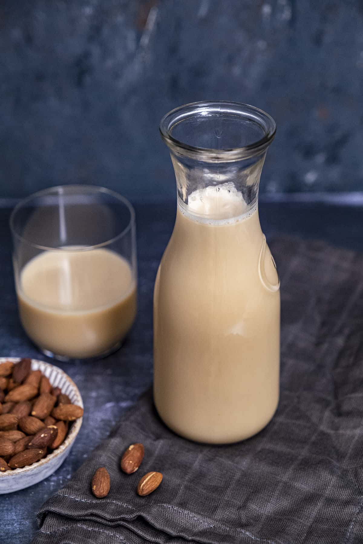 Homemade almond milk in a bottle and in a glass and roasted whole almonds on the side on a dark background.